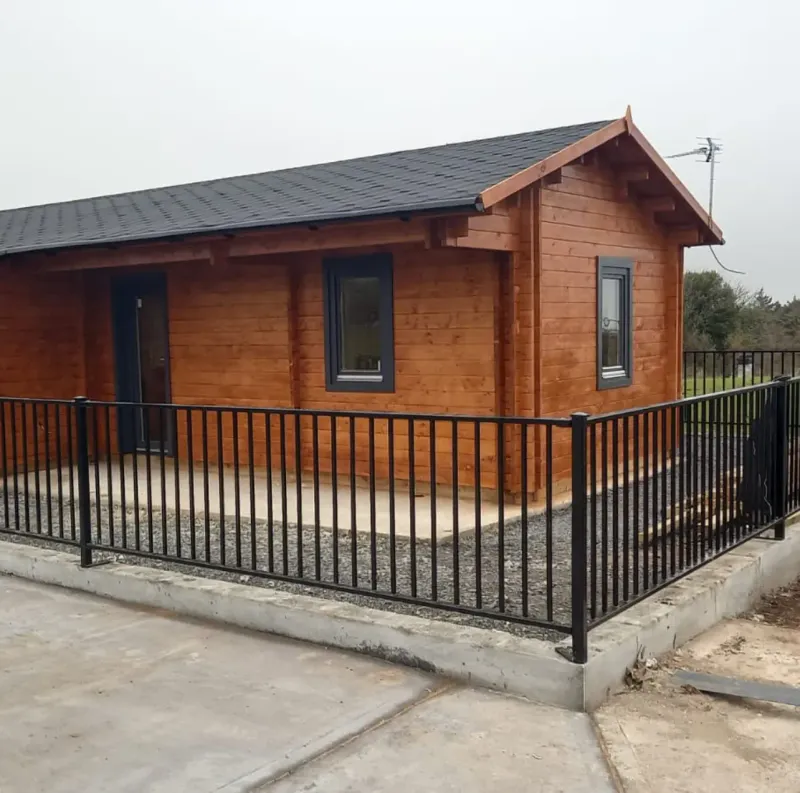 Small wooden cabin with dark gray roof and black metal fence surrounding the concrete patio.