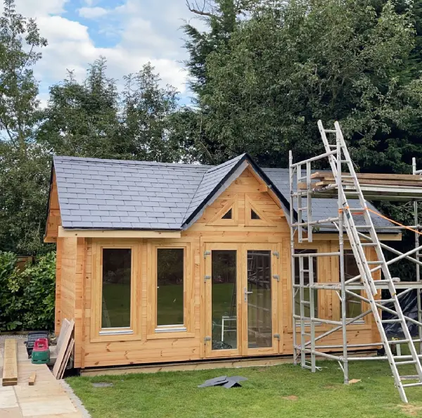 New wooden garden shed with slate roof under construction, surrounded by green lawn and trees, with scaffolding on the right side.