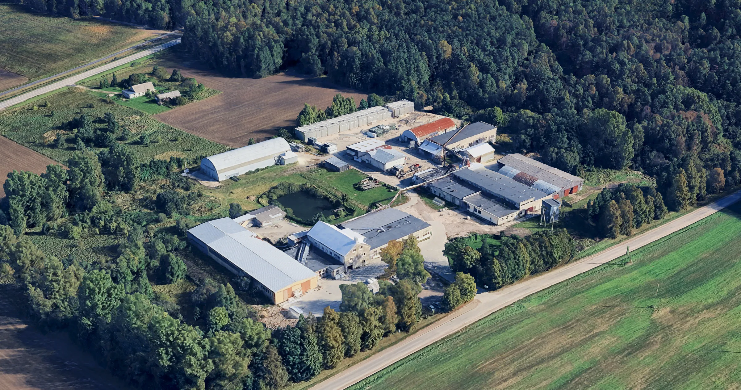 Aerial 3D view of the Satus Baltic factory in Vydmantai: production halls, timber yard and warehouses surrounded by forest and fields.